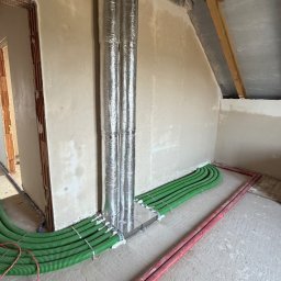 Dan-Kan Instalacje Grzewcze I Sanitarne Daniel Kania - Zielone rury instalacyjne ułożone wzdłuż ściany i pod sufitem, z pionowymi, izolowanymi przewodami. Surowy stan wykończenia wnętrza, widoczne elementy konstrukcji dachu.