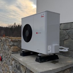 Dan-Kan Instalacje Grzewcze I Sanitarne Daniel Kania - Biała pompa ciepła Saunier Duval Genie Air Split zamontowana na kamiennym cokole przy ścianie budynku, widok z boku, zachodzące słońce w tle.