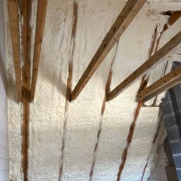 AlfaPur Ocieplenia Pianą Pur - Poddasze z drewnianą konstrukcją pokryte grubą warstwą białej pianki izolacyjnej. Widoczny fragment ściany i podłogi oraz urządzenie do natrysku pianki.