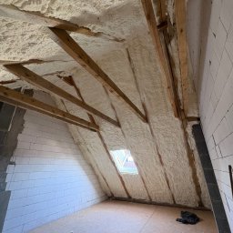 AlfaPur Ocieplenia Pianą Pur - Poddasze ocieplone pianką PUR, widoczne drewniane belki konstrukcyjne i fragment okna dachowego. Biała pianka pokrywa skośne ściany i sufit, tworząc jednolitą warstwę izolacyjną.