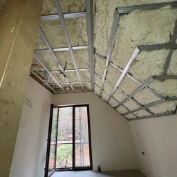 AlfaPur Ocieplenia Pianą Pur - Poddasze z widoczną konstrukcją metalową i wypełnieniem z pianki izolacyjnej, okno z widokiem na zewnątrz. Etap prac ociepleniowych w nowym domu.
