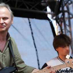 A0 Przemysław Schab - Muzyk grający na gitarze basowej podczas koncertu na scenie, widoczny mikrofon i drugi muzyk w tle. Ujęcie z bliska, skupione na gitarzyście.