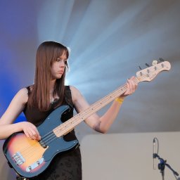 A0 Przemysław Schab - Młoda kobieta gra na gitarze basowej na scenie, oświetlona reflektorami. Widoczny mikrofon i fragment publiczności. Ujęcie z bliska, skupione na muzyku.