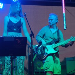 A0 Przemysław Schab - Zespół muzyczny na scenie podczas występu: wokalistka w zebrze, basista z gitarą, perkusista w okularach. Scena oświetlona niebieskim i kolorowym światłem.