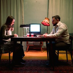 Studio Illusio - Dwoje ludzi siedzi przy stole w pokoju oświetlonym lampami i telewizorem. Na stole stoją szklanki z napojem. Scena przypomina kadr z filmu lub sesji zdjęciowej.