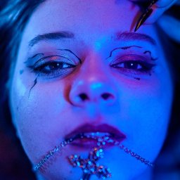 Studio Illusio - Portret kobiety z artystycznym makijażem oczu i srebrnym łańcuszkiem z krzyżem w ustach, oświetlony intensywnym niebieskim światłem. Dłoń z długimi, czarnymi paznokciami dotyka czoła.