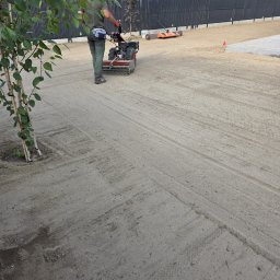 GARDENPOL - Pracownik w zielonym strooju obsługuje czerwoną glebogryzarkę, precyzyjnie przygotowując piaszczysty grunt pod trawnik. Widoczne młode drzewko i czarne ogrodzenie.