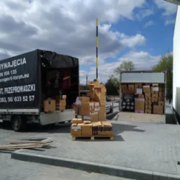 Dwa busy dostawcze, jeden z plandeką z napisem 'DO WYNAJĘCIA TRANSPORT, PRZEPROWADZKI' i numerami telefon&oacute;w, drugi otwarty, oba załadowane kartonami, na pierwszym planie paleta z kartonami, w tle...