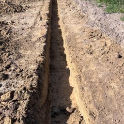 Wiktor Fajkowski - Wąski wykop pod fundament z widoczną linką wyznaczającą linię, obok zbrojenie i trawa. Ziemia o zróżnicowanej strukturze i kolorystyce. Słoneczny dzień.