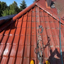 Alpinblue - Czerwona dachówka częściowo pokryta mchem, zabezpieczenia linowe, żółte buty pracownika. Widok z góry na dach podczas czyszczenia lub mycia.