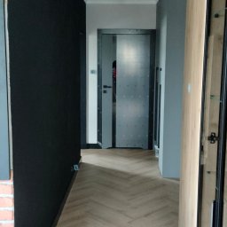 REMKOR Wykończenia pod klucz - Nowoczesny korytarz z parkietem w jodełkę, czarną ścianą z ceglanym akcentem i oświetleniem punktowym. Widok na metalowe drzwi i przeszkloną szafę.