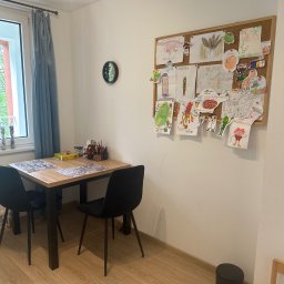 Ewelina Janiak-Wojdyńska psychoterapia i pomoc psychologiczna - Gabinet dziecięcy z drewnianym stołem, czarnymi krzesłami i tablicą korkową z rysunkami dziecięcymi. Okno z zasłoną i zegar na ścianie.
