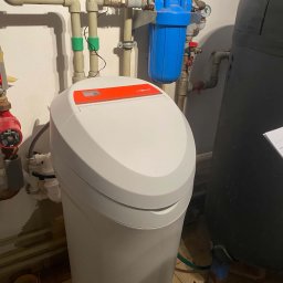 Kacper Gajof - Biały zmiękczacz wody Viessmann z pomarańczowym panelem, widoczne rury instalacji wodnej z filtrem i zaworami, oraz czarny zbiornik. Ujęcie z bliska.