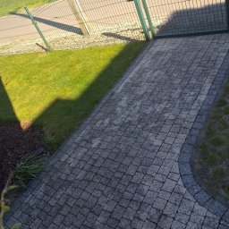 Das dekor - Ułożona kostka brukowa tworząca ścieżkę w ogrodzie, widoczna z perspektywy lekko z góry, z zielonym trawnikiem i fragmentem ogrodzenia w tle.