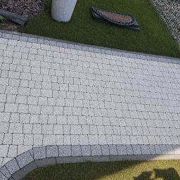 Das dekor - Jasnoszara kostka brukowa ułożona wzdłuż trawnika z elementami dekoracyjnymi, widok z góry. Obrzeża z ciemniejszej kostki. Fragment siatki ogrodzeniowej w tle.