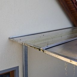 Das dekor - Czyszczenie zadaszenia nad wejściem do domu. Woda spływa z umytej powierzchni, ukazując efekt czyszczenia. Widoczna elewacja budynku i fragment dachu.