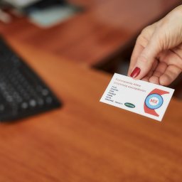 UBEZPIECZENIA MULTIAGENCJA MIX - Dłoń z czerwoną emalią prezentuje wizytówkę ubezpieczeniową MIX na biurku z klawiaturą. Oferta obejmuje życie, dom, podróż, firmę, auto i zdrowie.