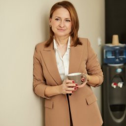 UBEZPIECZENIA MULTIAGENCJA MIX - Kobieta w beżowym garniturze, oparta o ścianę, trzyma szary kubek. W tle dystrybutor wody. Uśmiechnięta, z krótkimi, rudymi włosami i delikatną biżuterią.