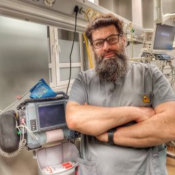 FIRST CARE - Medyk z brodą w szarym uniformie, ze skrzyżowanymi rękami, na tle sali z aparaturą medyczną, w tym defibrylator Lifepak 15 i monitor Mindray. Okulary na nosie.