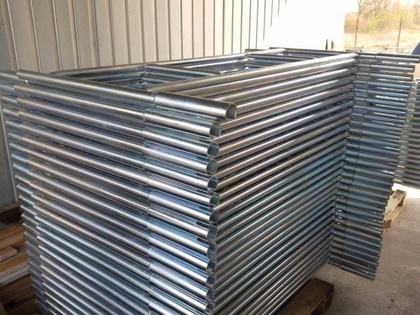 Stosy ocynkowanych, metalowych ram budowlanych na drewnianych paletach, widoczne w magazynie.