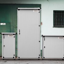 SICA - Trzy obudowy SICA HEATBOX na magazyny energii o różnych rozmiarach, ustawione na zewnątrz budynku z zielonymi drzwiami i oknem z kratą. Szare, metalowe konstrukcje.