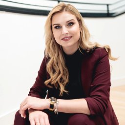 POSTRONIE Marta Badowska - Portret uśmiechniętej blondynki w bordowym garniturze, siedzącej na schodach. Zegarek na ręce, jasne tło, naturalne światło. Profesjonalny wizerunek.
