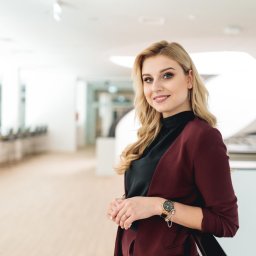 POSTRONIE Marta Badowska - Portret uśmiechniętej blondynki w bordowej marynarce, opierającej się o białą balustradę w nowoczesnym biurze. Zegarek na ręce, subtelny makijaż.