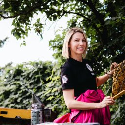 POSTRONIE Marta Badowska - Uśmiechnięta blondynka w czarnej koszulce prezentuje ramkę z pszczołami na tle zieleni, obok dymka pszczelarska. Ujęcie z lekkiej perspektywy.