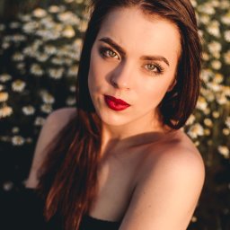 POSTRONIE Marta Badowska - Portret kobiety z intensywną czerwoną szminką na tle kwitnącej łąki. Ujęcie z góry, podkreślające makijaż i naturalne światło.