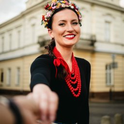 POSTRONIE Marta Badowska - Uśmiechnięta kobieta w ludowym stroju z kwiatowym wiankiem i czerwonymi koralami, wyciąga dłoń w stronę obiektywu na tle jasnego budynku.