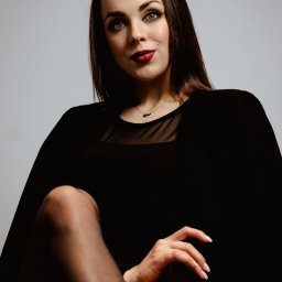 POSTRONIE Marta Badowska - Portret kobiety w czarnej sukience z siateczkowym dekoltem i bordową szminką, spoglądającej z góry, z nogą założoną na nogę i dłonią w kadrze.