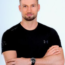 Michał Trzonkowski - Mężczyzna w czarnej koszulce sportowej Under Armour, z zegarkiem na lewym nadgarstku, stoi na jasnym tle z założonymi rękami. Krótkie włosy i zadbana broda.