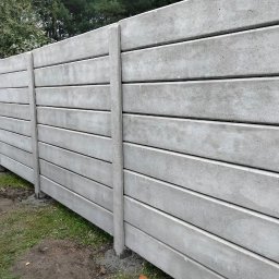 JDG OLEKSANDR VITVITSKYI - Szary, prefabrykowany mur z poziomych betonowych paneli, oddzielonych pionowymi słupkami, na tle zielonej trawy i drzew. Widok z boku, lekko z dołu.