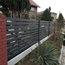JDG OLEKSANDR VITVITSKYI - Nowoczesne, poziome przęsła w grafitowym odcieniu, osadzone na niskiej, betonowej podmurówce i ceglanym słupku. W tle domy i zieleń.