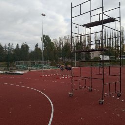 JDG OLEKSANDR VITVITSKYI - Boisko sportowe z czerwoną nawierzchnią, częściowo ogrodzone siatką. Widoczne rusztowanie na kółkach i elementy wyposażenia sportowego. Pochmurne niebo w tle.