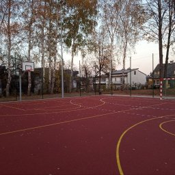 JDG OLEKSANDR VITVITSKYI - Ogrodzenie boiska sportowego z bramką do piłki nożnej i koszem do koszykówki, otoczone drzewami. Czerwona nawierzchnia boiska z żółtymi liniami.
