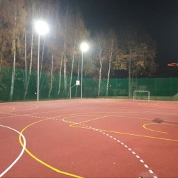 JDG OLEKSANDR VITVITSKYI - Oświetlony nocą, wielofunkcyjny plac sportowy z czerwoną nawierzchnią, zielonym ogrodzeniem z siatki i drzewami w tle. Widoczne bramki i kosze do koszykówki.