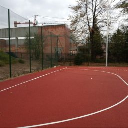 JDG OLEKSANDR VITVITSKYI - Czerwone boisko sportowe z siatką ochronną i bramką w tle, widok na budynek w budowie z dźwigiem. Pochmurne niebo.