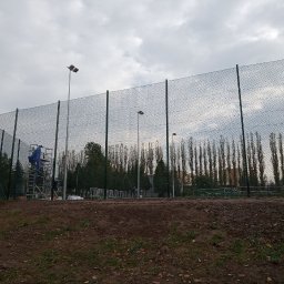 JDG OLEKSANDR VITVITSKYI - Montaż siatki ogrodzeniowej na boisku sportowym. Widoczny pracownik na rusztowaniu, wysokie słupy oświetleniowe i rząd drzew w tle. Pochmurne niebo.