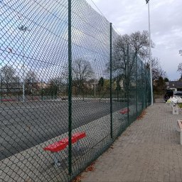 JDG OLEKSANDR VITVITSKYI - Zabezpieczenie boiska siatką ogrodzeniową w zielonej ramie. Widoczne ławki i chodnik z kostki. W tle zabudowa jednorodzinna i drzewa.