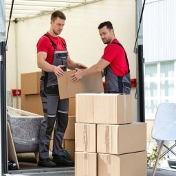 HMMM LOGISTICS Sp. z o.o. - Dwóch mężczyzn w kombinezonach układa kartony wewnątrz furgonetki. W tle budynek z białymi ścianami i oknami. Widok z tyłu pojazdu.