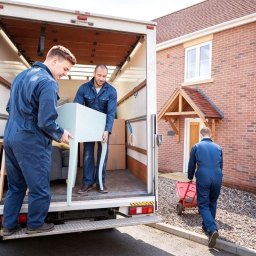 HMMM LOGISTICS Sp. z o.o. - Dwóch mężczyzn w kombinezonach wnosi jasnoniebieski stolik do furgonetki przeprowadzkowej. Trzeci mężczyzna z wózkiem idzie w stronę domu z cegły.