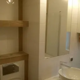 Nowoczesna łazienka z białymi płytkami, drewnianymi półkami, okrągłą umywalką nablatową i kulistymi lampami wiszącymi, widok na kabinę prysznicową w lustrze.