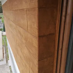 Detal elewacji domu z brązową okładziną imitującą drewno, widoczny narożnik i fragment okna z drewnianą ramą, obok rusztowanie.