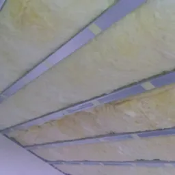 Montaż wełny mineralnej w metalowym stelażu pod sufitem, zabezpieczonej taśmą, widok z dołu na skośną powierzchnię.