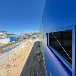 Hydrobros 777 - Nietypowy kadr na budowę hali z niebieską elewacją z blachy trapezowej i otworem okiennym. Widoczne przygotowanie terenu i elementy konstrukcyjne.