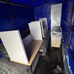 Przeprowadzki STRONGERSI - Wnętrze pojazdu transportowego z zabezpieczonymi meblami: szara sofa, białe półki z drewnianymi blatami i szafka owinięta folią. Niebieska plandeka i metalowa konstrukcja.