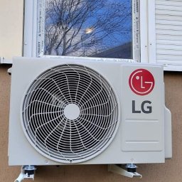 Vortex-montujemy komfort - Zamontowana jednostka zewnętrzna klimatyzacji LG na ścianie budynku, widoczna pod oknem z roletami. Na szybie okna odbija się korona drzewa.