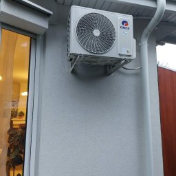 Vortex-montujemy komfort - Biała jednostka zewnętrzna klimatyzacji marki Gree zamontowana na ścianie budynku, widoczna rynna i okno z roślinami w tle. Ujęcie z dołu.