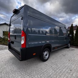 Jakub Rudowski - Szary bus dostawczy z widocznym tyłem i bokiem, zaparkowany na kostce brukowej. W tle budynek i pochmurne niebo. Widoczne detale: relingi dachowe, stalowe felgi.